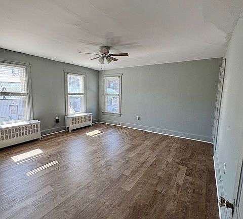 Bright and inviting bedroom with warm wood-look flooring and soft gray walls, creating a modern yet cozy atmosphere. A ceiling fan with overhead lighting provides comfort and energy efficiency year-round. Natural light streams in from the window.