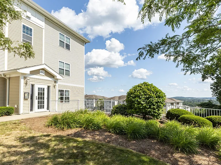 Exterior view of apartment building with beige siding and lush landscaping with view of additional buildings and panoramic view in background at Summit Place apartments in Methuen, MA.