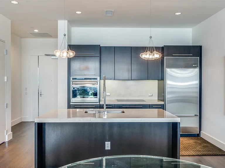 Kitchen featuring stainless steel appliances, pendant lighting, a center island with sink, dark wood-type flooring, and light stone counters