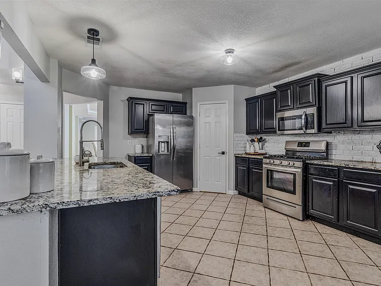 Kitchen featuring granite-style countertops, dark cabinetry, stainless steel appliances, a white brick-style backsplash, and neutral tile flooring