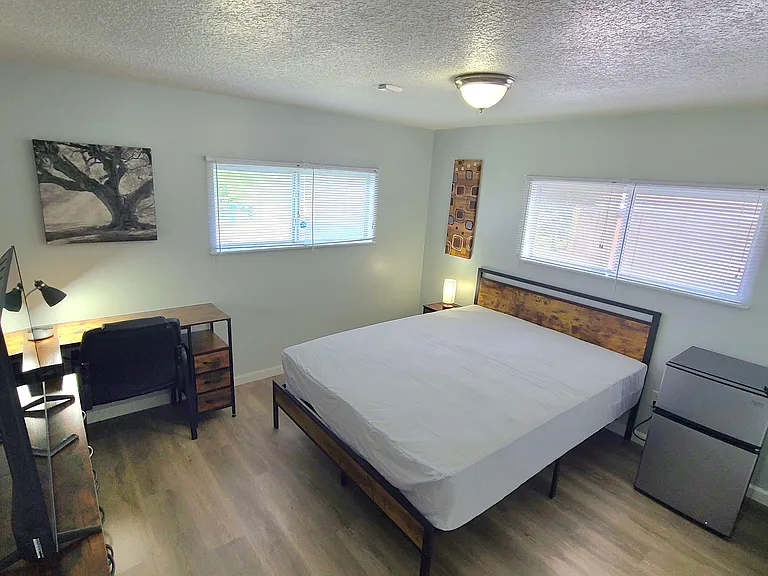 Furnished bedroom with a full bed, work desk, TV, and mini fridge, plus two windows with blinds for natural light.
