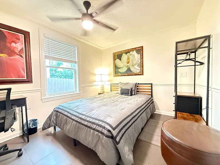Furnished bedroom featuring a ceiling fan, window with blinds, and space for a desk plus open hanging/storage and Ottoman