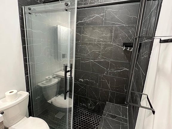 Spacious, beautifully tiled bathroom featuring porcelain surfaces throughout, a large walk-in shower with built-in bench seating, a sleek frameless glass sliding door, and both a rainfall and handheld shower head for a luxurious experience.