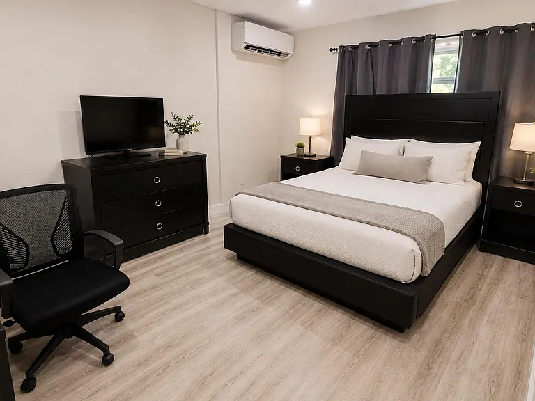 Furnished bedroom with a queen bed, nightstands, dresser with TV, and a dedicated desk area plus split AC and window.