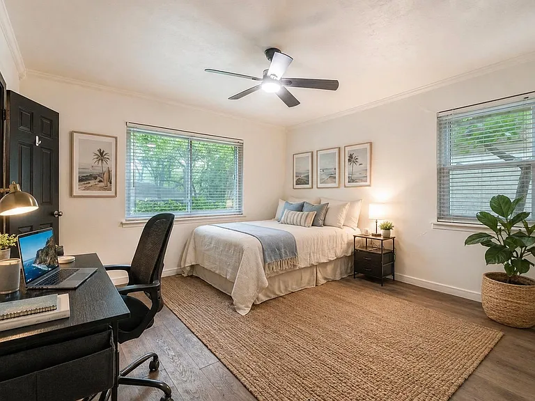 Spacious bedroom with desk workspace, ceiling fan, and large windows with blinds plus a nightstand and soft area rug.