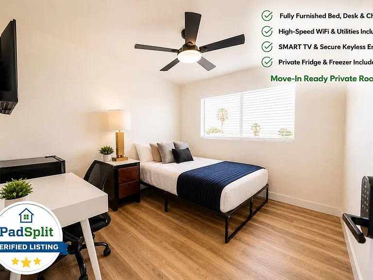 Furnished private bedroom with desk setup, wall-mounted TV, ceiling fan, and a window for natural light.