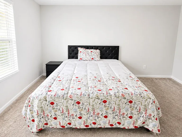 Comfortable bedroom with a full/queen bed, nightstand, carpet flooring, and a large window with blinds for natural light.