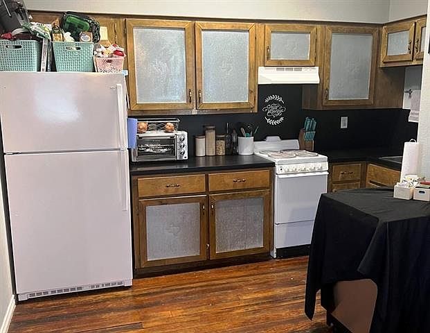 Kitchen with white appliances, dark wood finished floors, dark countertops, custom exhaust hood, and glass insert cabinets