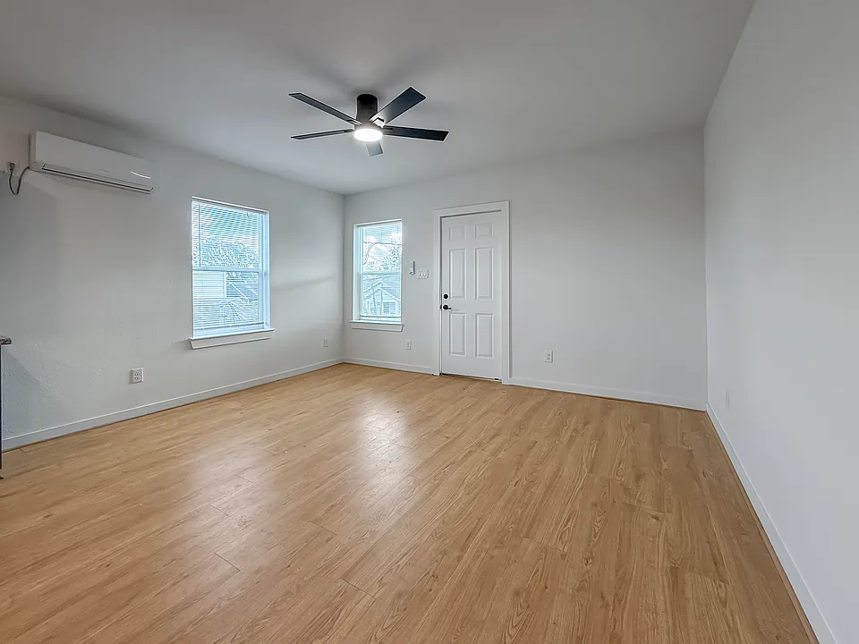 Bright, spacious room with light wood flooring, white walls, and a ceiling fan. Features a mini-split AC unit and two windows for natural light. Ideal for a bedroom or office space.