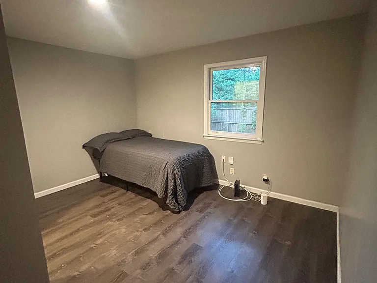 Bedroom with wood-look flooring, neutral walls, and a large window overlooking the yard; ample space for a full bed setup.