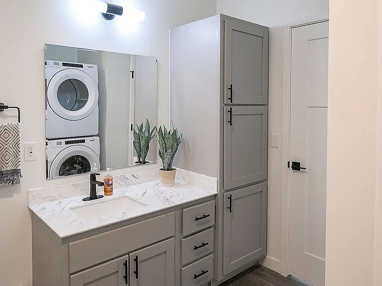 Modern bathroom with sleek gray cabinetry, marble countertop, and in-unit stacked washer and dryer for convenience.