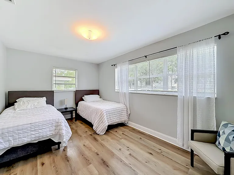 Spacious bedroom with two twin beds, wood-look flooring, and large windows with blinds and curtains for natural light.