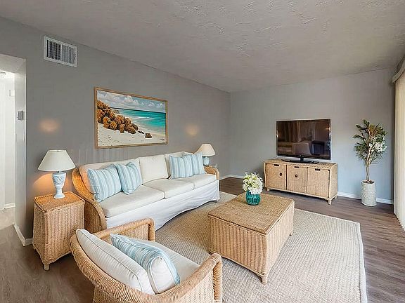 Inviting living room with elegant wicker furniture, a cozy white couch, and a widescreen TV, complemented by soothing beach-themed artwork and abundant natural light.