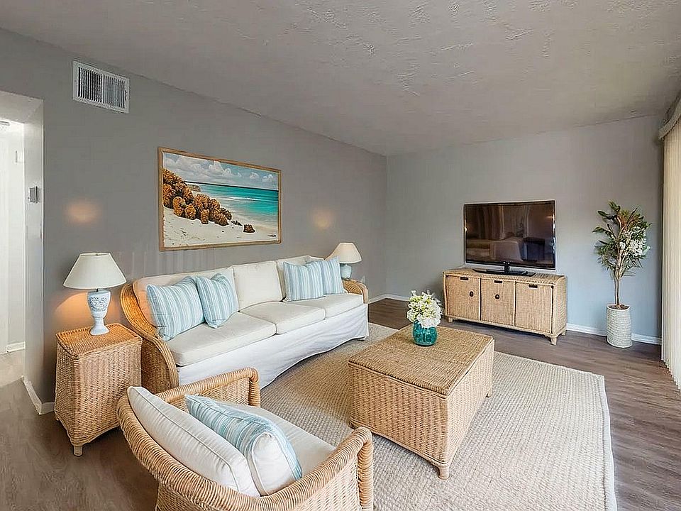 Inviting living room with elegant wicker furniture, a cozy white couch, and a widescreen TV, complemented by soothing beach-themed artwork and abundant natural light.