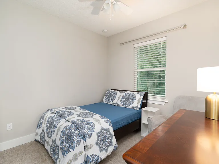 This serene bedroom setup features a built-in ceiling fan and large windows framing quiet, green outdoor views