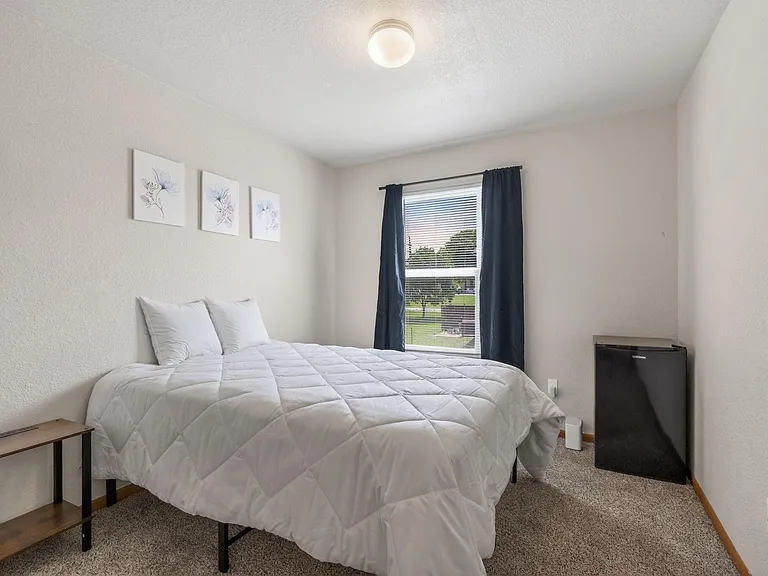 Private bedroom with a full-size bed, window with curtains, and space for a dresser or desk; includes a mini fridge.