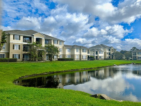 Charming apartment complex with elegant architecture, lush green landscape, and a serene pond under a vibrant blue sky, offering a picturesque living environment.