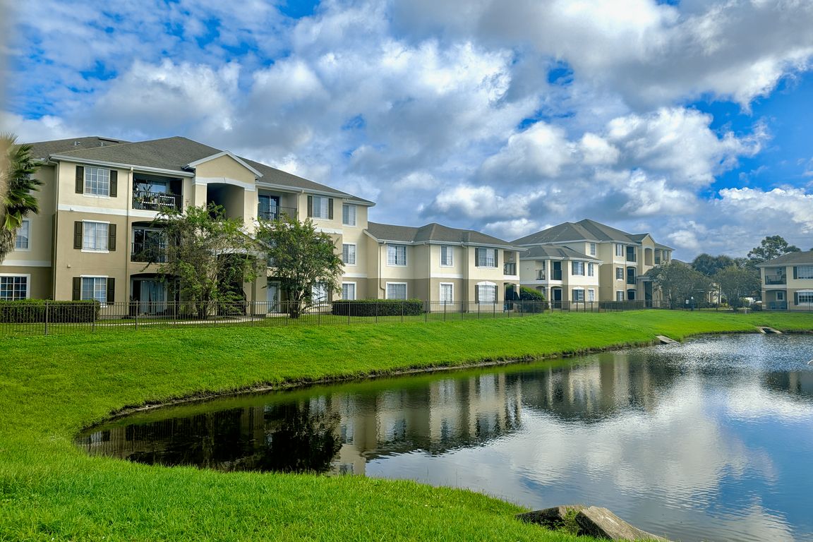 Charming apartment complex with elegant architecture, lush green landscape, and a serene pond under a vibrant blue sky, offering a picturesque living environment.
