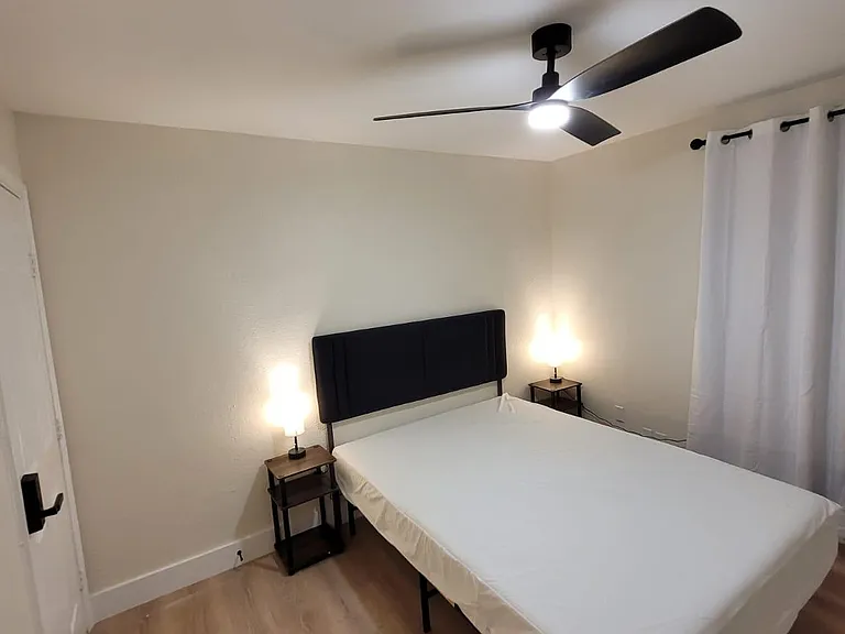 Furnished bedroom with a comfortable bed, matching nightstands and lamps, ceiling fan, and window with curtain panels.