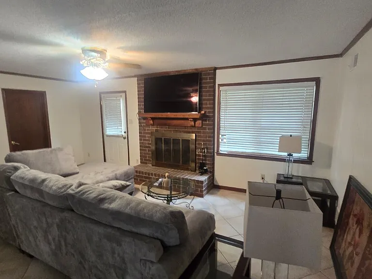 Comfortable living room with a brick fireplace, mounted TV, large window blinds, and generous seating space.