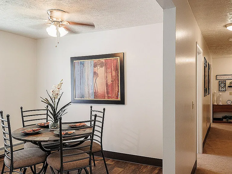 Charming dining area with modern decor, featuring a round table set for four under a stylish ceiling fan, adjacent to a bright hallway adorned with elegant artwork.