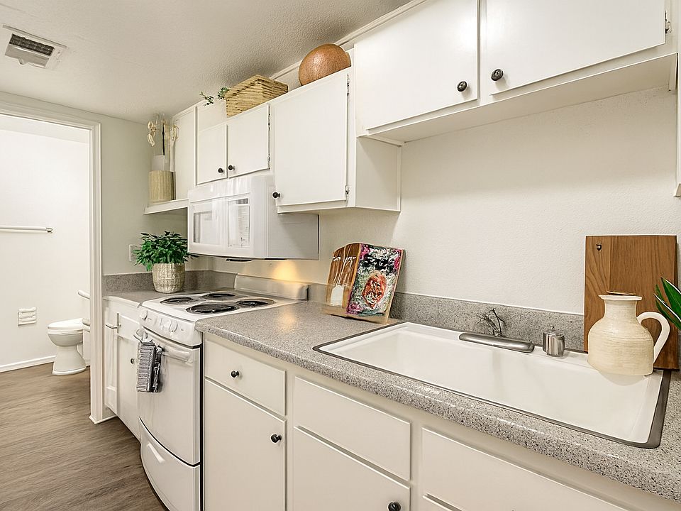 Bright galley kitchen with white cabinetry, speckled countertops, and dark wood-look flooring. Offers ample storage, modern appliances, and a clean layout with space for cooking essentials and personal touches. Convenient access to adjacent bath.