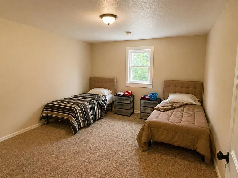 Spacious carpeted bedroom with two twin beds, a centered window, and room for dressers or a small desk setup.