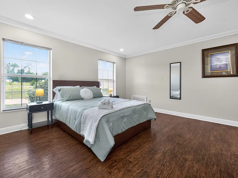 Spacious bedroom with hardwood floors, ceiling fan, and two windows for natural light, plus bedside table and mirror.