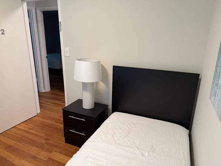 Furnished bedroom with wood floors, a bed with headboard, and a nightstand with lamp, plus easy access to the hallway.