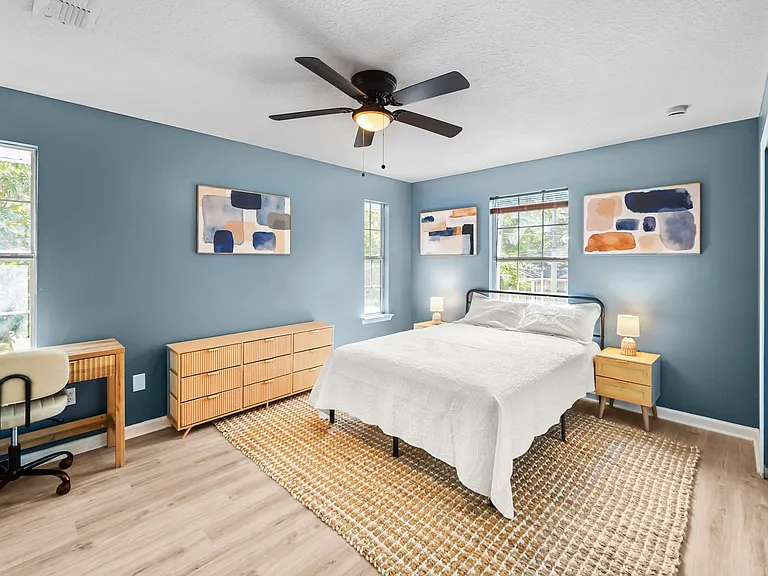 Spacious bedroom with multiple windows, ceiling fan, work desk, and ample storage with dresser and nightstands.