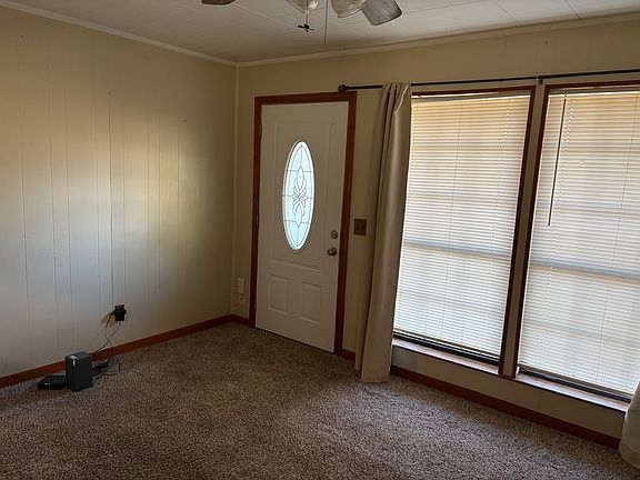 Living room with large window, decorative front door, carpet & ceiling fan.