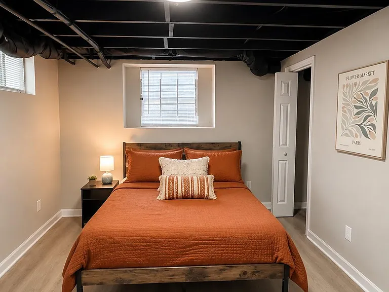 Staged bedroom with a full/queen bed, nightstand lighting, and a window with blinds on durable wood-look flooring.
