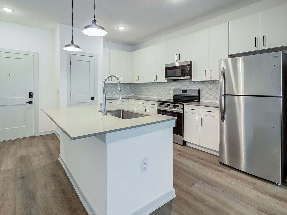 White shaker cabinets, a kitchen island with seating, glass-top stove, built-in microwave, and stainless steel refrigerator _ finished with wood-style plank flooring and designer pendant lighting.