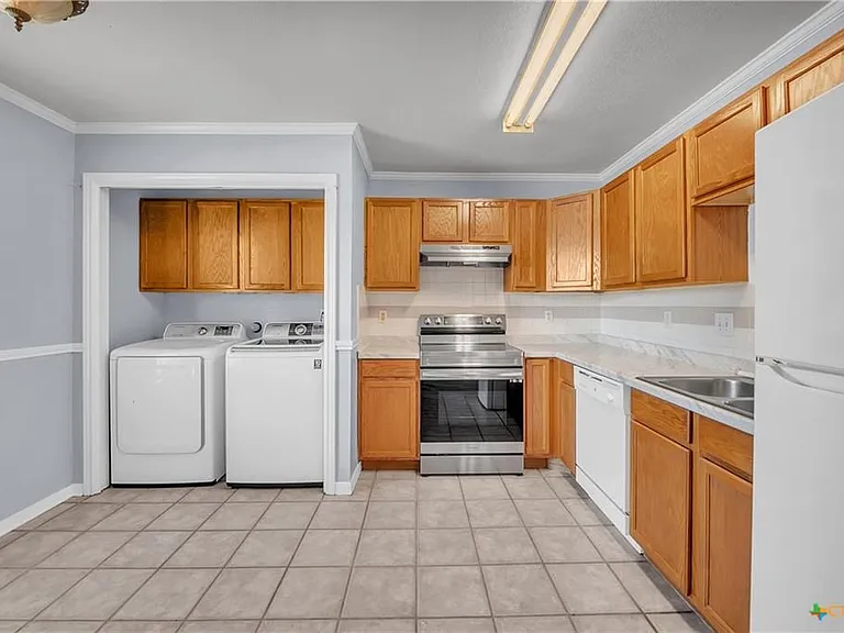 A kitchen space separated from the living room with a washer and dryer, stove, oven, and range, garbage disposal, dishwasher and fridge. The cabinets have been painted since this photo.