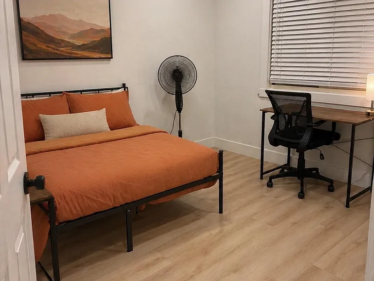 Furnished bedroom with a full bed, dedicated desk workspace, and window with blinds over wood-style flooring.
