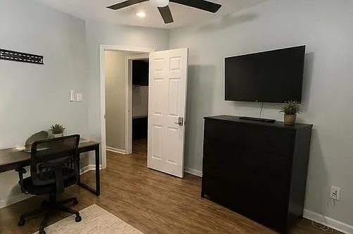 Private bedroom with ceiling fan, hardwood-style floors, and a dedicated desk area plus dresser and wall-mounted TV.