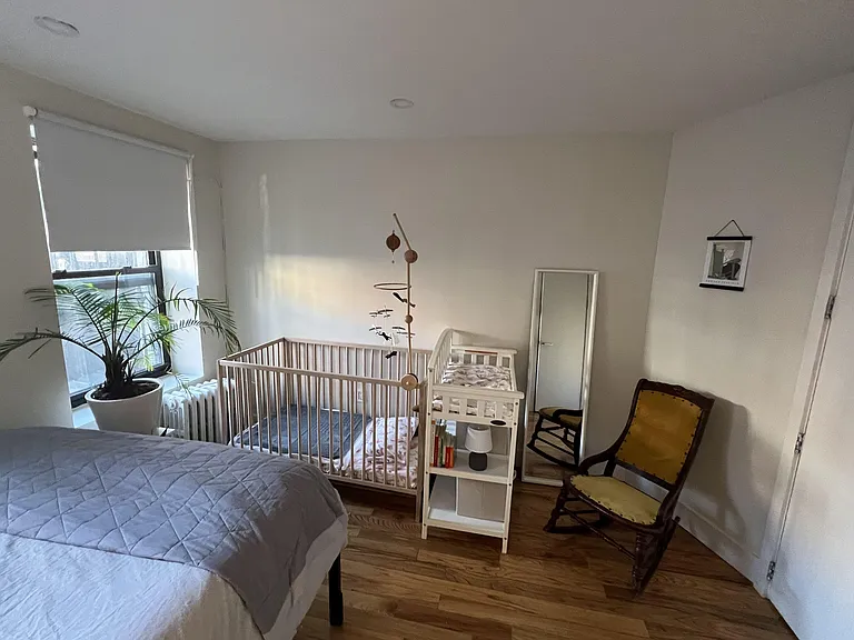 Bedroom with large window, wood floors, and space for a bed plus seating, with a freestanding mirror for added function.