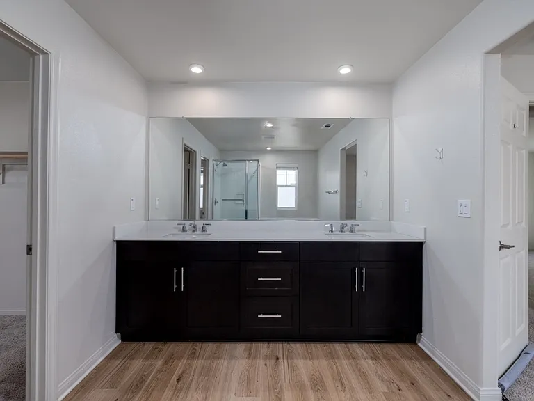 Stylish master bathroom featuring dual sinks, a large wall-to-wall mirror, and elegant laminate flooring. Modern lighting and sleek dark cabinetry create a bright, luxurious space perfect for daily comfort and convenience.