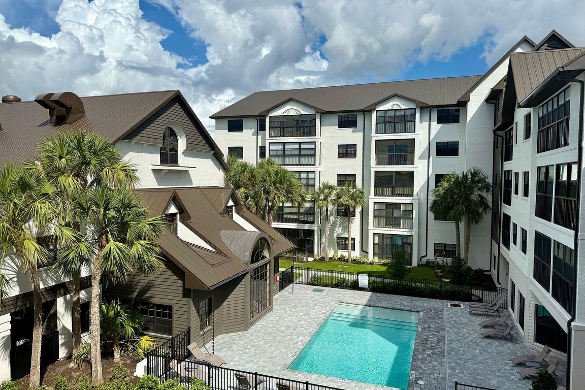 Inviting apartment complex with a serene swimming pool, surrounded by lush palm trees and modern architecture under a vibrant sky.