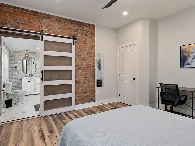 Bedroom with exposed brick accent wall, barn-style door, and a dedicated desk area with easy access to an adjacent bath.