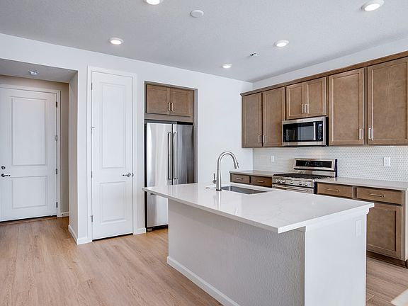 A modern, open-concept kitchen with light wood cabinets, white countertops, and stainless steel appl