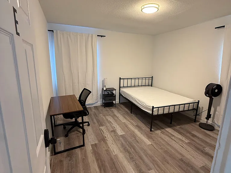 Private furnished bedroom with a work desk, nightstand, and durable wood-look flooring plus window coverings.