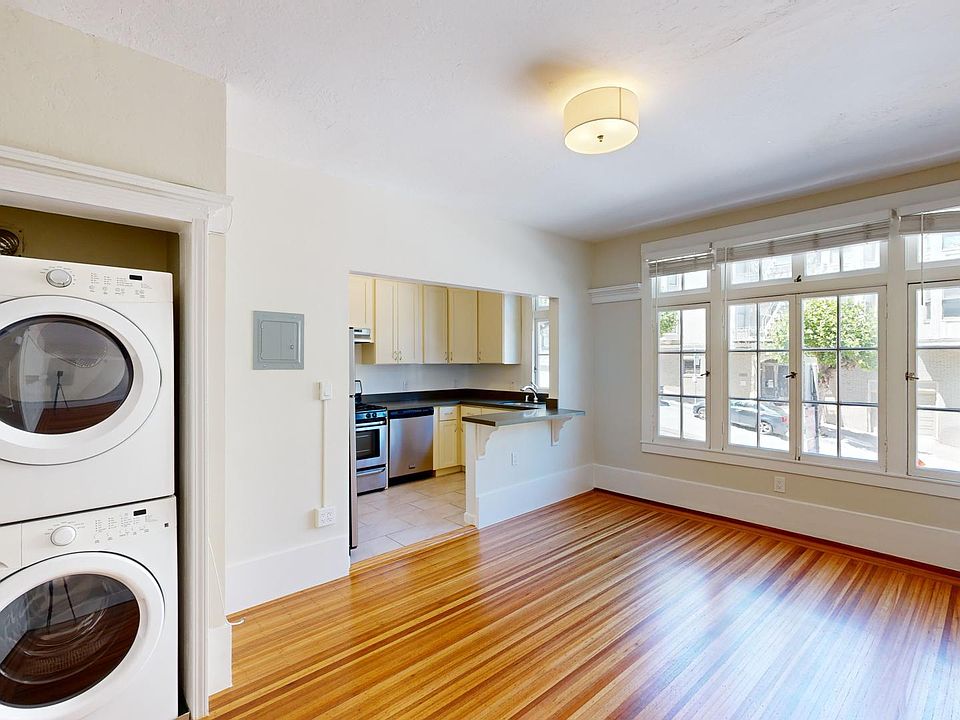 Bright laundry area with stacked washer and dryer, open to a kitchen featuring cabinets, and a spacious living area with hardwood floors.