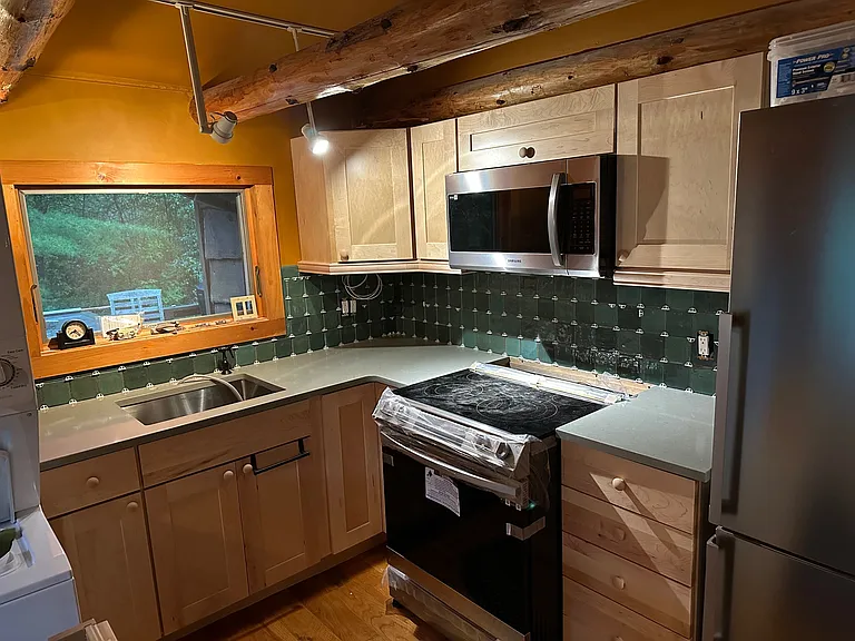 Kitchen Area with sink and window, quartz countertops, hanging cabinets, electric stove, microwave and refrigerator with forest green tile backsplash.