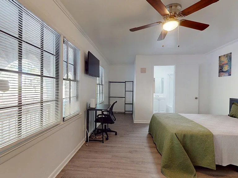 Spacious bedroom with large windows, ceiling fan, wall-mounted TV, and a dedicated desk area near the ensuite bath.