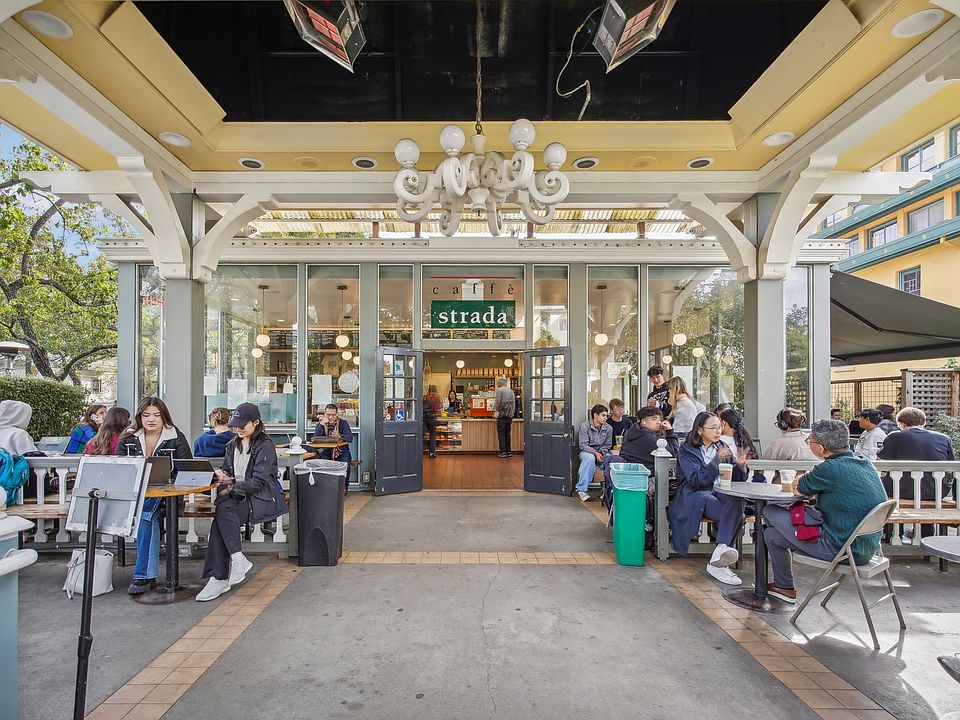 Bustling outdoor cafe with a large glass entrance framed by columns, filled with people seated at tables