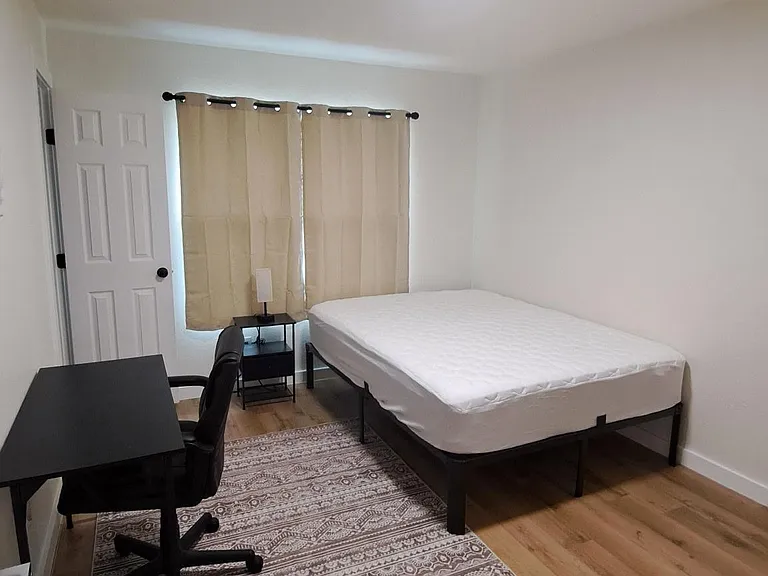 Furnished bedroom with a full bed, desk workspace, and nightstand, plus wood-look flooring and a window with curtains.