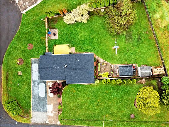 Overhead view of entire lot showing off parking on the left front, fruit trees & small garden patch on side yard, back deck with firepit nearby, raised flowerbed and garden to the right of the house, with shop next to that riding lawnmower that stays between outbuildings, storage shed further right and stacked rows of cut wood for the fireplace at the far right.