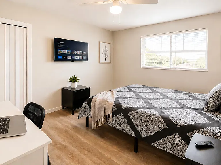 Furnished bedroom with a full bed, work desk, wall-mounted TV, and a sliding-door closet plus a large window.
