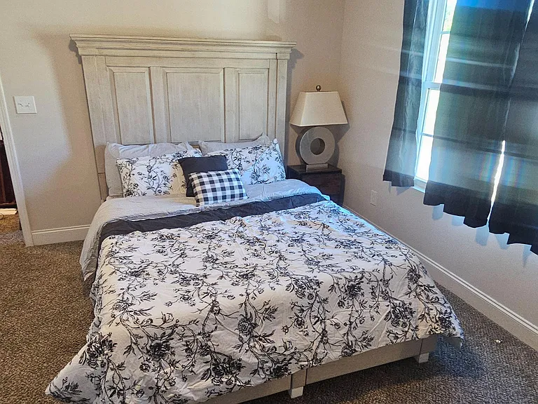 Furnished bedroom with a full-size bed, nightstand and lamp, plus a window with curtains and soft carpet underfoot.